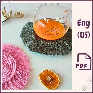May include: A clear glass mug with the words "Good Morning" in black script sits on a green macrame coaster. A pink macrame coaster and a dried orange slice are also visible. The mug contains an orange liquid.