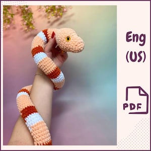 May include: A crocheted snake toy with orange and white stripes. The snake is curled up and has a light pink body. The toy is being held by a hand.