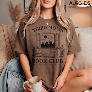 Può includere: T-shirt marrone con la scritta "TIRED MOMS BOOK CLUB" e una grafica di una finestra che si affaccia sullo skyline della città. La maglietta presenta anche la scritta "RUNNING ON COFFEE, CHAOS AND CHAPTERS".