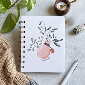 May include: A white spiral-bound notebook with a graphic illustration of a pink bottle containing leafy branches, a small tag with a crescent moon, and the text "FOOD JOURNAL" printed in black at the bottom center of the cover.