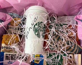 Puede incluir: Una caja de regalo llena de una taza de café Starbucks con el nombre "Vanya" escrito, bocadillos y paquetes de café Starbucks. La caja está forrada con papel de seda rosa y hebras de papel blanco.