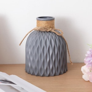 May include: A grey vase with a textured, wavy pattern. The vase is decorated with natural rope tied around the neck. The vase is on a wooden surface.