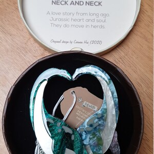 May include: A pair of green and white dinosaur-shaped earrings in a round box. The earrings are made of resin and have a glittery finish. The box has a black interior and a white label with the text "Neck and Neck" and "Original design by Carmen Hui (2021)".