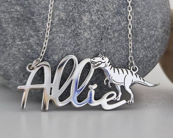 Puede incluir: Collar de plata con el nombre "Allie" en cursiva, acompañado de una silueta de dinosaurio. El colgante cuelga de una cadena delicada, sobre un fondo neutro.