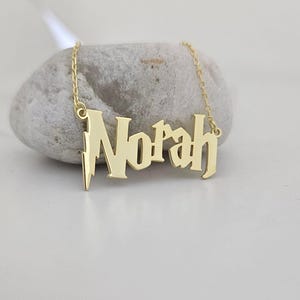 Puede incluir: Un collar dorado con el nombre "Norah" en una fuente estilizada. El colgante está suspendido de una delicada cadena. El collar se muestra sobre una piedra gris sobre un fondo blanco. El diseño de rayo está en el lado izquierdo del nombre.