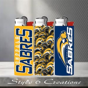 K&ouml;nnte beinhalten: Drei Feuerzeuge mit unterschiedlichen Designs. Eines ist gelb mit "BUFFALO SABRES" in Blau, ein anderes hat ein Muster aus gelben und schwarzen S&auml;bel-Logos, und das dritte ist blau mit dem Teamlogo und "BUFFALO SABRES".