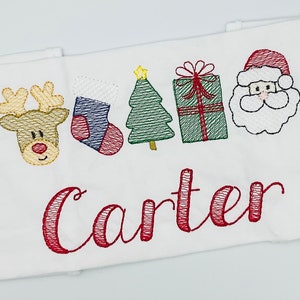 以下が含まれることがあります： 白い生地に刺繍されたクリスマスデザイン。トナカイ、靴下、木、プレゼント、サンタクロースが含まれています。「Carter」という名前が赤い筆記体で刺繍されています。全体的なテーマは、お祝いと休日に関連しています。