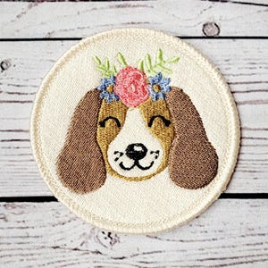 Peut inclure: Patch brodé représentant un chien brun et blanc portant une couronne de fleurs. Le chien a une expression joyeuse et regarde directement le spectateur.