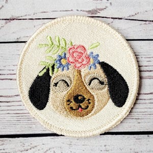 May include: Embroidered patch of a dog wearing a flower crown. The dog has brown fur, black ears, and a pink tongue. The flowers are pink, blue, and green.