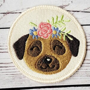 Pode incluir: Etiqueta bordada de um pug marrom usando uma coroa de flores com flores rosa, azuis e amarelas. A etiqueta é circular e tem um fundo branco.