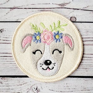 May include: Embroidered patch of a white dog with pink ears and a flower crown. The dog has a black nose and a pink tongue. The flowers in the crown are pink, blue, and yellow.