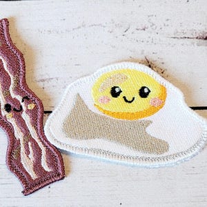 May include: Two felt patches, one shaped like a piece of bacon and the other like a fried egg. Both have smiling faces with eyes and a mouth.