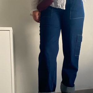 Op de afbeelding: Een persoon draagt een wit linnen shirt met een bruine leren tas en donkerblauwe jeans met witte stiksels. Ze dragen bruine leren enkellaarsjes met veters.