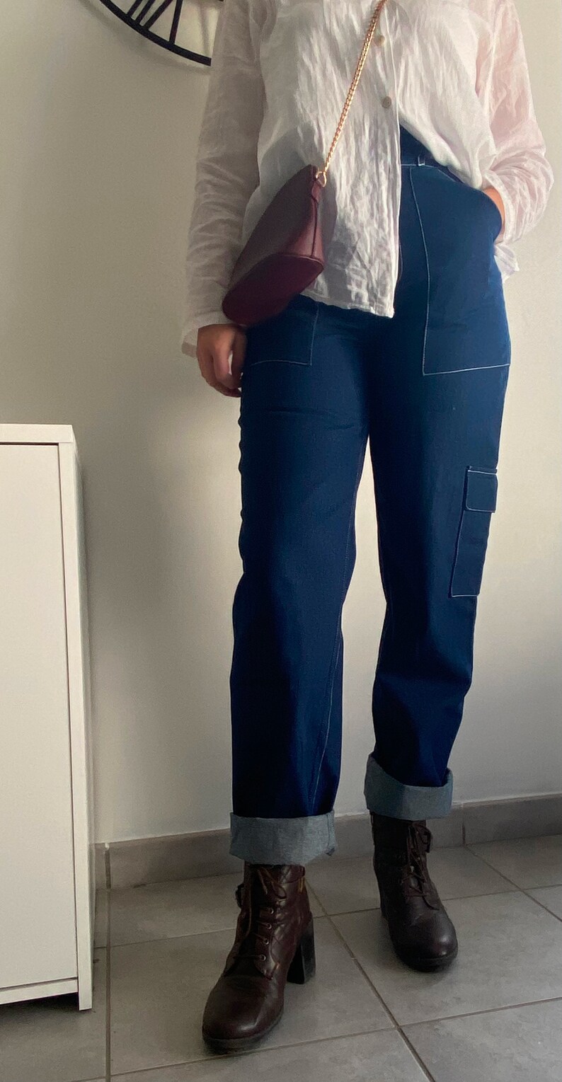 Op de afbeelding: Een persoon draagt een wit linnen shirt met een bruine leren tas en donkerblauwe jeans met witte stiksels. Ze dragen bruine leren enkellaarsjes met veters.