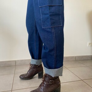 Op de afbeelding: Donkerblauwe denim cargo broek met een grote cargotas op het rechterbeen. De broek is omgeslagen aan de onderkant en de persoon draagt ​​bruine enkellaarsjes.
