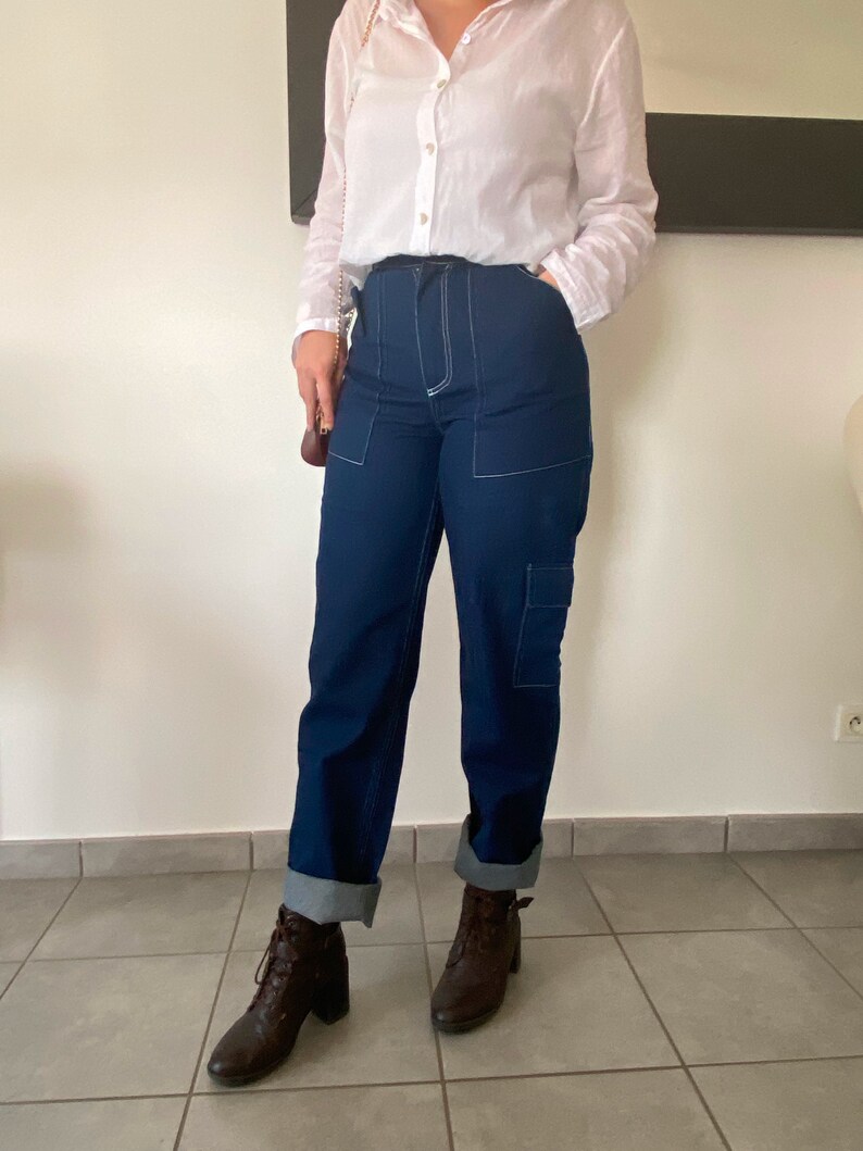 Op de afbeelding: Een vrouw draagt een wit overhemd met knopen, een donkerblauwe jeans met witte stiksels en bruine enkellaarsjes. De jeans heeft grote zakken aan de zijkanten.
