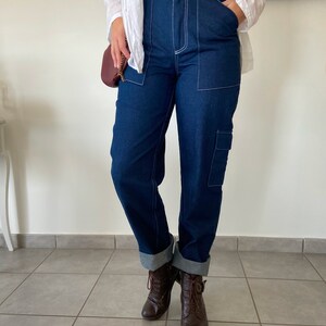 Op de afbeelding: Een donkerblauwe spijkerbroek met witte stiksels. De broek heeft een hoge taille en een rechte pijp. De broek is aan de onderkant omgeslagen en wordt gedragen met bruine veterschoenen.