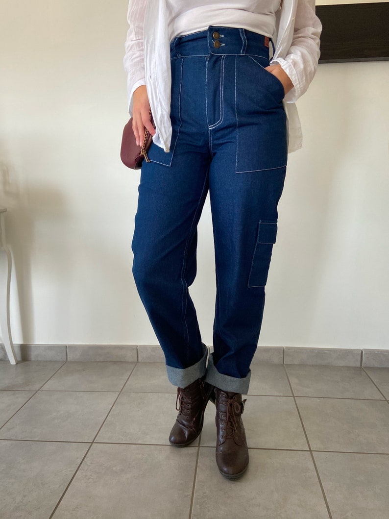 Op de afbeelding: Een donkerblauwe spijkerbroek met witte stiksels. De broek heeft een hoge taille en een rechte pijp. De broek is aan de onderkant omgeslagen en wordt gedragen met bruine veterschoenen.