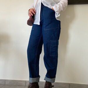 Op de afbeelding: Een vrouw draagt een wit overhemd met knopen en een gouden ketting, donkerblauwe jeans met witte stiksels en bruine enkellaarsjes.