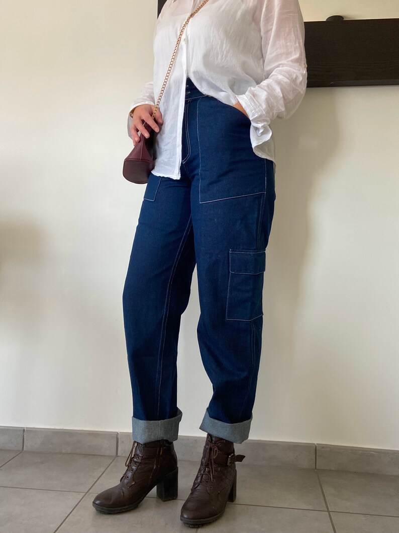 Op de afbeelding: Een vrouw draagt een wit overhemd met knopen en een gouden ketting, donkerblauwe jeans met witte stiksels en bruine enkellaarsjes.