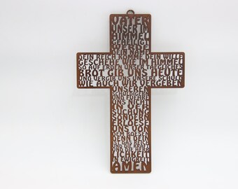May include: A rustic metal cross with the Lord's Prayer cut out in the shape of the cross. The cross is a warm brown color and hangs against a white background. The text is in German.
