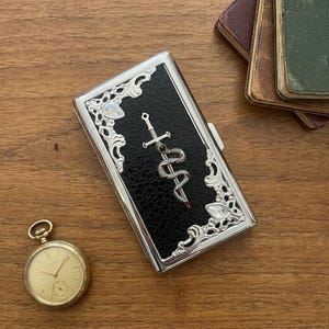 以下が含まれることがあります： 黒いテクスチャーの長方形のシガレットケース。四隅には白い石が埋め込まれた銀色の装飾的なフィリグリーが施されています。中央には、蛇が巻き付いた剣の銀色のエンブレムがあり、剣の柄には小さな赤い石があしらわれています。