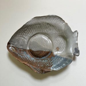 May include: A handmade ceramic fish-shaped plate with a central recessed area. The plate features a color palette of gray, brown, and blue, with textured details mimicking fish scales. The plate is designed for serving or display.