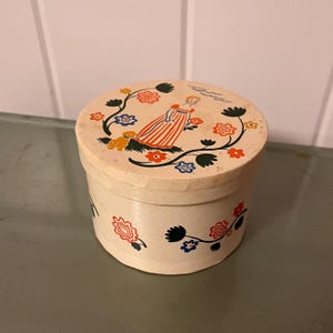 May include: A round, cream-colored box with a lid featuring a vintage illustration. The lid depicts a woman in a striped dress with a dog, surrounded by floral designs in red, blue, and yellow. The box sides have similar floral patterns.