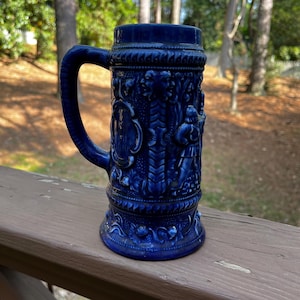 May include: A cobalt blue beer stein with a handle. The stein is decorated with relief carvings of figures, faces, and floral patterns. The base and rim have detailed designs. The stein is approximately 8 inches tall.