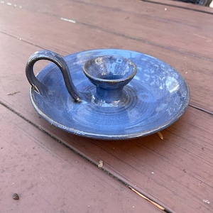 May include: A handmade blue ceramic candle holder with a handle. The candle holder has a circular base with a raised center for the candle and a curved handle. The ceramic has a glossy finish.
