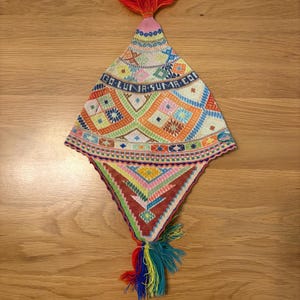 May include: A colorful, hand-stitched hat with geometric patterns and the text "GO LUNA SUMA CO". The hat features a red tassel at the top and a rainbow tassel at the bottom. The hat is displayed on a wooden surface.