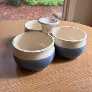 May include: A set of three white ceramic bowls with blue rims, each with a separate compartment. The bowls are arranged in a circle around a central white ceramic spout.