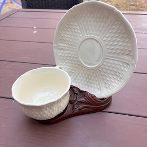 May include: A white porcelain teacup and saucer with a raised, textured design. The cup and saucer are displayed on a wooden stand.