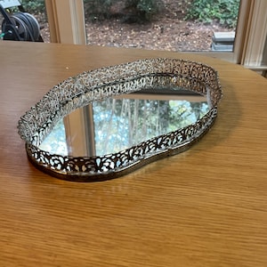May include: An oval-shaped vanity tray featuring a reflective mirrored base and a decorative silver-toned metal gallery rail. The raised edge displays an intricate, openwork filigree pattern that encircles the entire perimeter of the tray.
