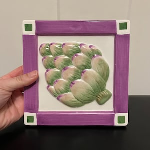 May include: A ceramic tile with a purple frame and green accents features a detailed, three-dimensional image of an artichoke.