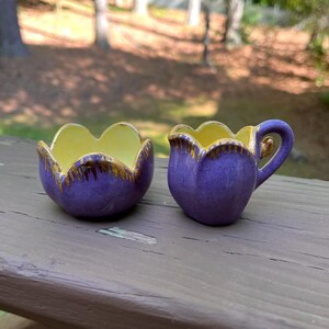 May include: A purple ceramic sugar bowl and creamer set. The interior of each piece is yellow, with a scalloped rim and gold trim. The creamer has a handle and a spout.