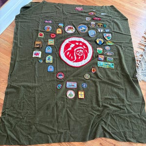 May include: An olive green blanket covered with a collection of colorful patches. The patches feature various designs, including badges, logos, and illustrations. A large red and white emblem is in the center. The blanket is on a wooden floor.