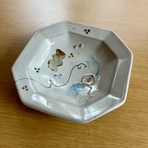 May include: A white ceramic octagonal serving dish with a hand-painted design in brown, blue, and red. The design features abstract shapes and lines.