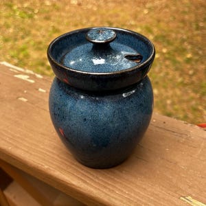 May include: A handcrafted, blue ceramic jar with a lid. The jar has a round body and a slightly flared rim. The lid sits on top and has a small knob. The jar has a speckled texture and a glossy finish. The jar is likely used for storing food or other items.