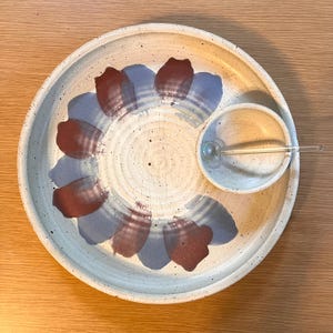 May include: A speckled ceramic serving platter with a built-in dip bowl and a clear glass stirrer. The platter is off-white with a floral design in shades of burgundy and blue. The dip bowl is also off-white.