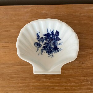 May include: A white porcelain seashell-shaped dish with a blue floral design. The dish features a central rose with surrounding leaves and smaller flowers.