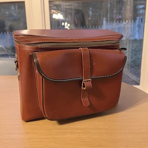 May include: A brown leather camera bag with a top zipper and a front flap secured by a strap and buckle. The bag has a rectangular shape with a side pocket and white stitching.