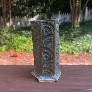 May include: A teal and brown ceramic vase with a geometric shape and a textured surface. The vase has a unique design with a repeating pattern of curved lines.