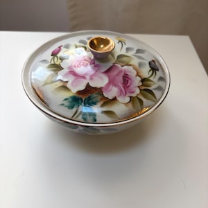 May include: A white ceramic bowl with a lid. The lid features a gold knob and is decorated with pink roses, green leaves, and gold trim. The bowl has a gold trim around the rim.