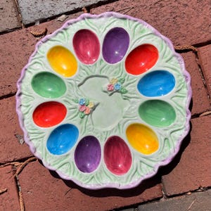 May include: A green and purple ceramic egg plate with 12 individual egg-shaped depressions. The plate is decorated with a bunny rabbit design and floral accents.