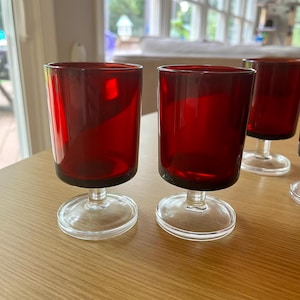 May include: Set of four red glass stemmed drinking glasses with clear glass bases. The glasses are on a wooden surface.