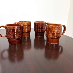 May include: A white ceramic mug with a black handle and the text "But First, Coffee" in a bold, black font. The mug is set against a blurred background, suggesting a cozy setting.