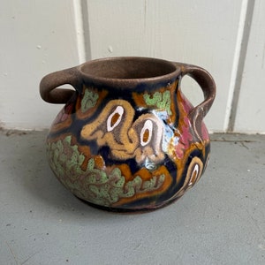 May include: A small, brown ceramic vase with two handles. The vase is decorated with a colorful floral pattern in shades of green, yellow, pink, and white.