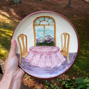 May include: A ceramic plate with a hand-painted scene of a table set with a pink tablecloth and two chairs, all in front of a window with a view of mountains. The plate has a brown rim.