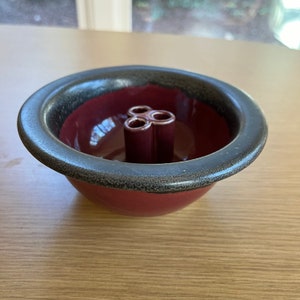 May include: A burgundy ceramic flower frog with a dark gray rim. The frog has a circular shape with a central stem and three holes for arranging flowers. The bowl is on a light brown surface.